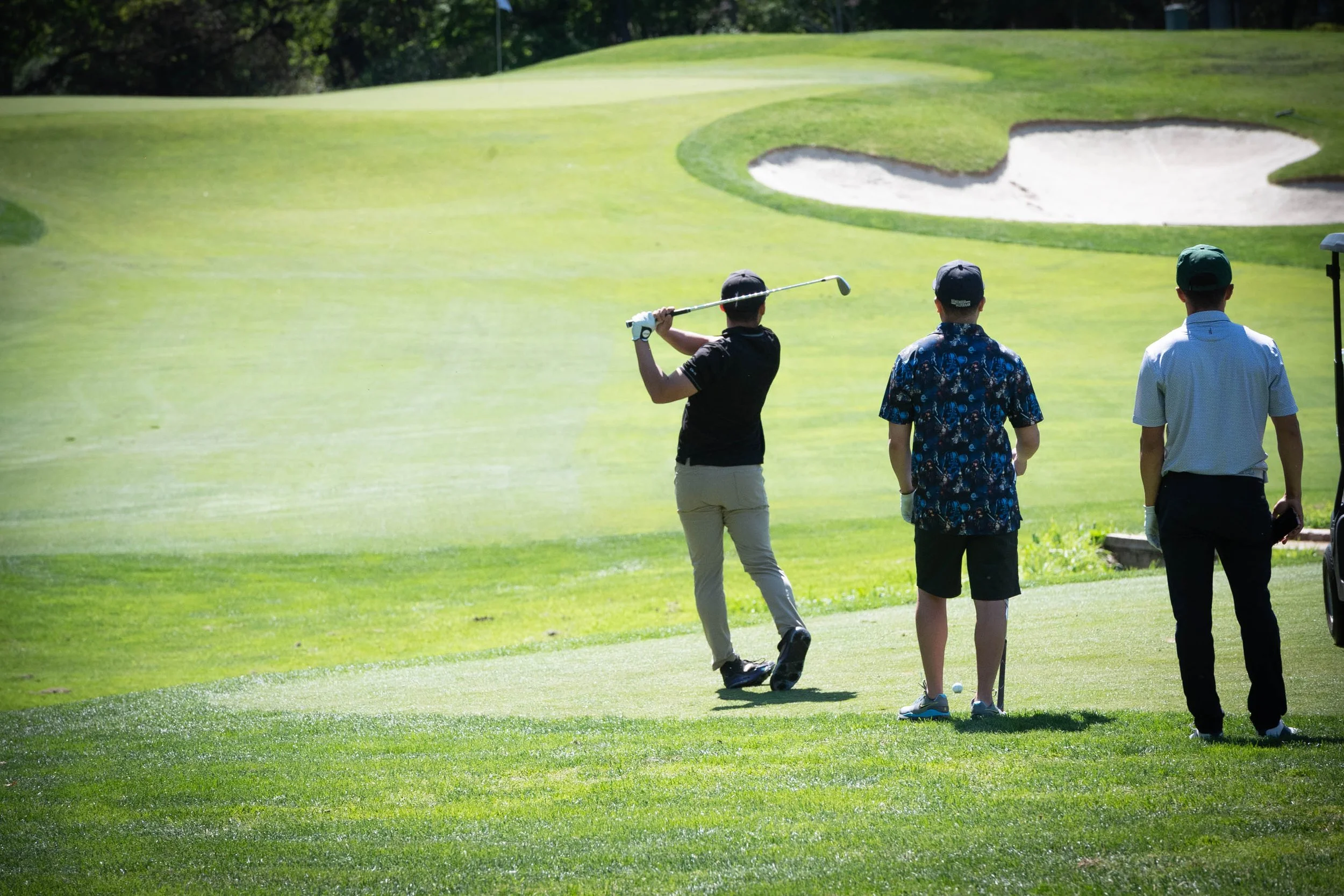 Three people on a golf course with one person about to hit a golf ball towards the green, while the other two watch.