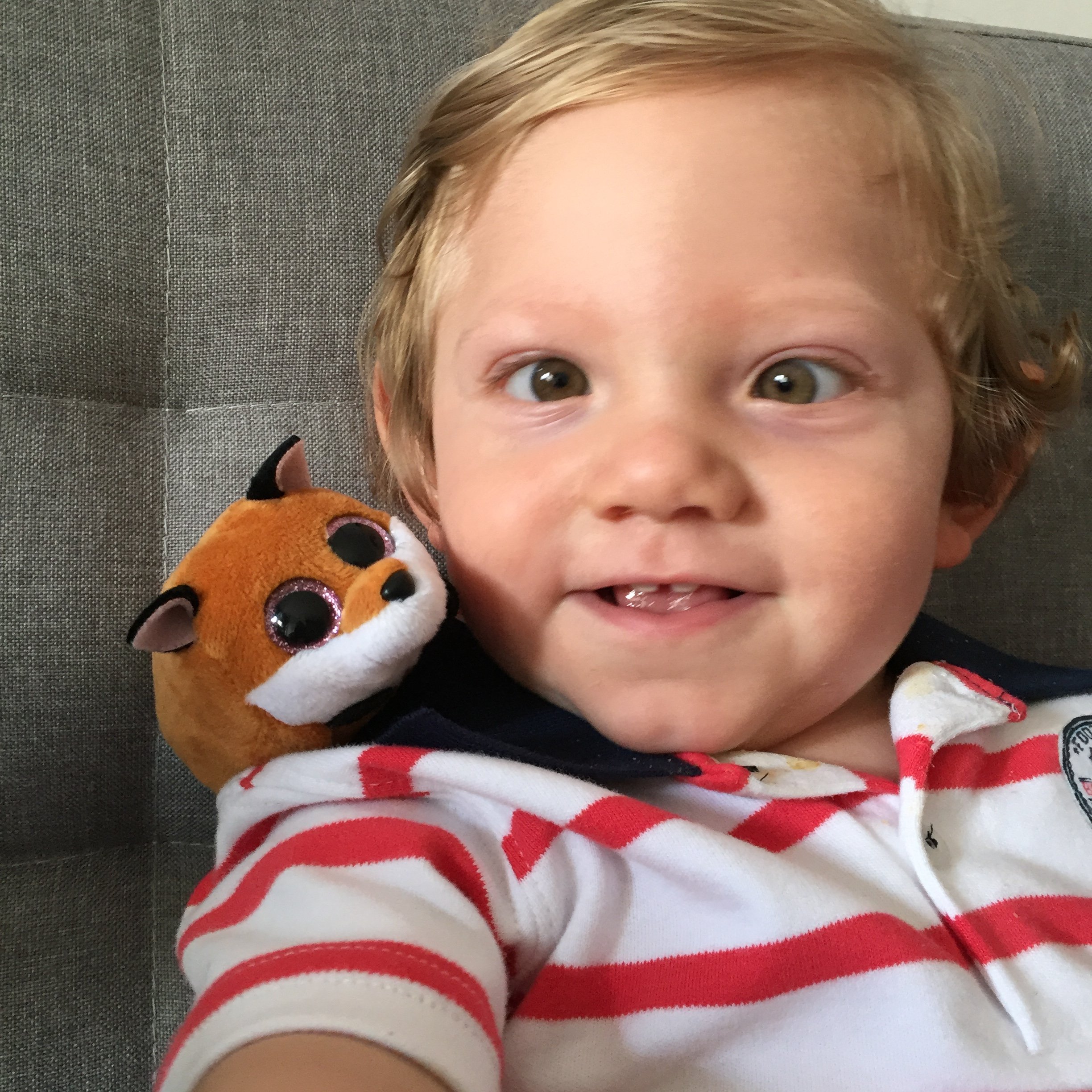 A young child with blonde hair and big brown eyes smiling, wearing a red and white striped shirt, sitting on a gray chair with a plush toy resting on their shoulder.