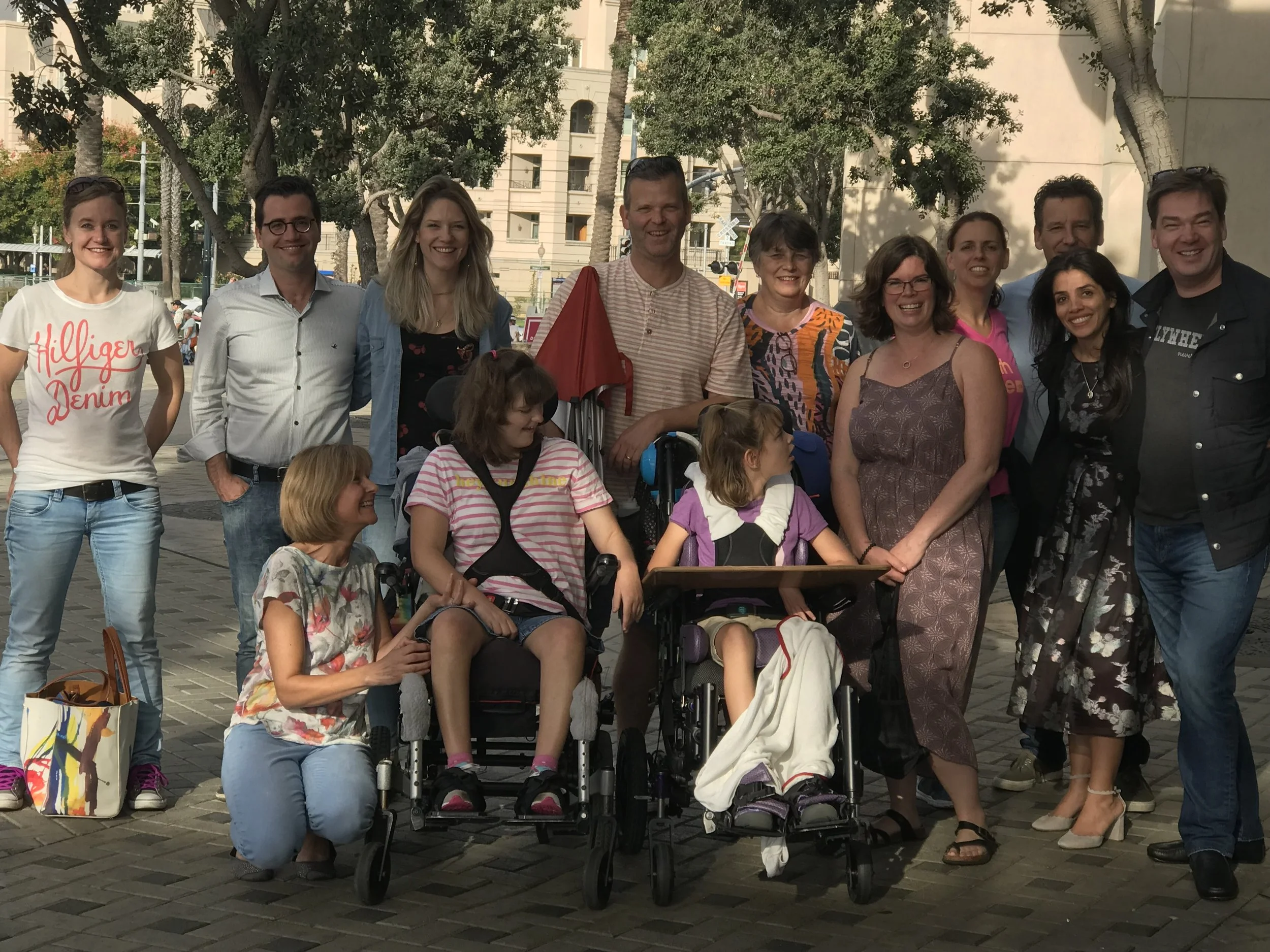 Group of people standing and sitting outdoors on a city street, with trees and tall buildings in the background. The group includes adults and children, some in wheelchairs, smiling and posing for the photo.